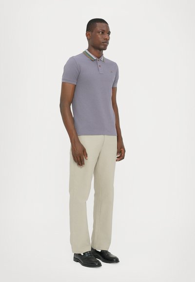Camisa polo de algodón lavanda con un cuello de rayas multicolores, combinada con pantalones beige y zapatos negros. El modelo se presenta de perfil.