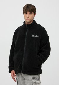 Czarna polarowa kurtka z zamkiem na całej długości i wysokim kołnierzem, z haftowanym napisem "dzika strona" na lewej piersi. Miękka, teksturowana powierzchnia, luźny krój.