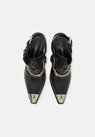 Black leather shoes with pointed toes, metallic silver accents, and a silver chain detail on the ankle strap. Flat soles for stability.