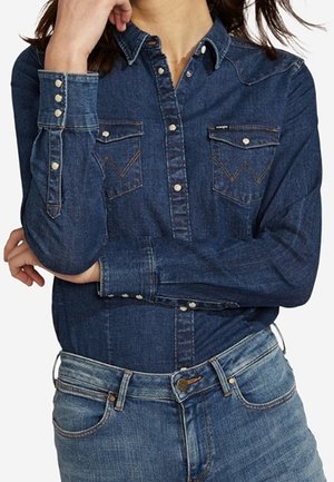 Personne portant une chemise en denim foncé à manches longues avec des boutons-pression et un jean bleu, un bras croisé et l'autre main près du visage.