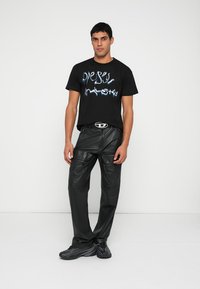 Crna majica s plavim grafičkim tekstom, uparena s crnim kožnim hlačama s više džepova i glatkom teksturom. Elegantne crne tenisice.