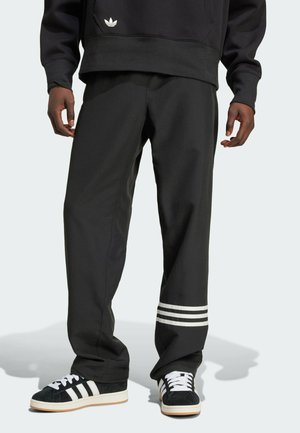 Sorte Adidas sweatpants med hvide tre-stribe accenter ved kanten, løs pasform, lavet af et glat stof, parret med sorte og hvide sneakers.