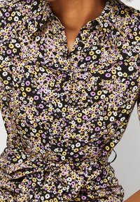 Chemise florale avec un fond noir ornée de fleurs blanches, jaunes et violettes. Design à boutonnière avec une taille cintrée et des manches courtes.