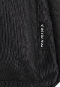 Konec černého texturovaného látkového batohu s bílou značkovou etiketou "Converse". Obsahuje zesílené švy a odolný zip.
