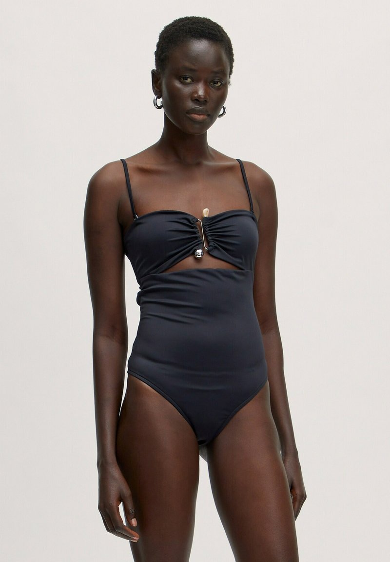 Zwarte badpak met dunne bandjes, voorzien van een gerimpelde voorkant en uitsnijding, verrijkt met een metalen hardware-element.