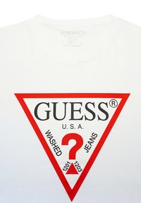 Weißes Baumwoll-T-Shirt mit einem roten dreieckigen Logo, auf dem die Wörter "GUESS", "WASHED", "JEANS" und ein Fragezeichen in der Mitte zu sehen sind.