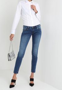 Camisa blanca de botones, jeans ajustados de mezclilla azul, tacones abiertos negros, sosteniendo un pequeño bolso de mano con un patrón en blanco y negro.