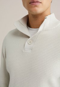 Svijetlosmeđi rastezljivi pulover s visokim ovratnikom, koji ima jedan gumb za zatvaranje. Mekani materijal s uzorkom u obliku rešetke. Nositi preko bijele košulje.