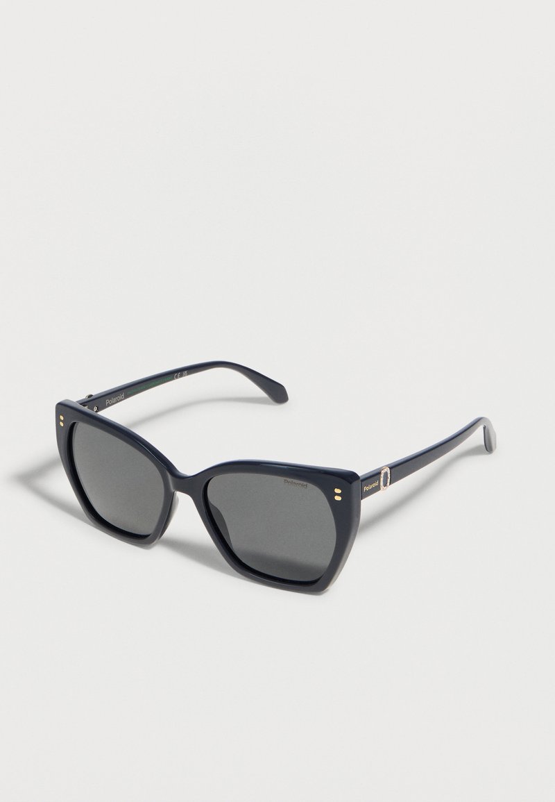 Lunettes de soleil polarisées à monture carrée noire avec verres foncés, accents dorés sur la charnière, et branches fines posées sur une surface blanche.