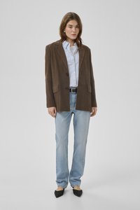 Vrouw staat, draagt een bruine blazer over een gestreept shirt met lichtblauwe jeans en zwarte puntige schoenen op een effen grijze achtergrond.