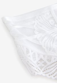 Lencería blanca de encaje con un delicado patrón que incluye secciones transparentes y tirantes elásticos, mostrando detalles intrincados y textura.