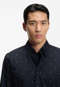 Camicia blu navy con bottoni, piccoli motivi a triangolo bianchi, colletto classico e maniche lunghe. Tessuto liscio, design aderente e leggero splendore.