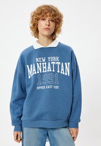 Blauwe sweatshirt met witte lettering "NEW YORK MANHATTAN 1991 UPPER EAST SIDE," met een witte kraag en een losse pasvorm.