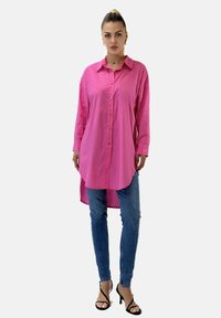 Rosa Langarmhemd mit Kragen und geschwungenem Saum. Kombiniert mit blauen skinny Jeans und schwarzen Riemchen-High Heels.