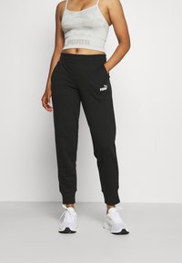 Pantalones deportivos negros con puños, que presentan un logo blanco de Puma. Combinados con un top corto de camuflaje gris claro y zapatillas deportivas blancas.