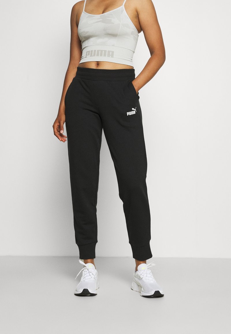 Pantalones deportivos negros con puños, que presentan un logo blanco de Puma. Combinados con un top corto de camuflaje gris claro y zapatillas deportivas blancas.