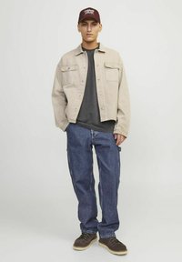 Jachetă din denim bej, cămașă gri dedesubt, blugi băieți albaștri, pantofi maronii și șapcă vișinie cu text alb. Outfit simplu, modern, casual.