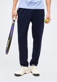 Άτομο που φοράει μπλε σκούρο παντελόνι και άσπρα αθλητικά παπούτσια κρατάει μια ρακέτα τένις στο ένα χέρι και μια μπάλα τένις στο άλλο.