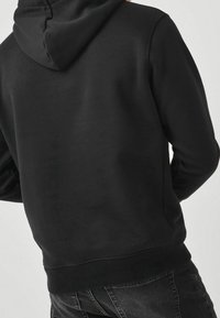 Vista posterior de una persona que lleva una sudadera negra lisa y jeans oscuros de lavado negro, contra un fondo claro.