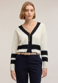 Kremowo-granatowy dzianinowy kardigan z wzorem warkoczowym, dekoltem w serek, paskami i zapięciem na guziki, zestawiony z granatowymi spodniami i beżowym paskiem.