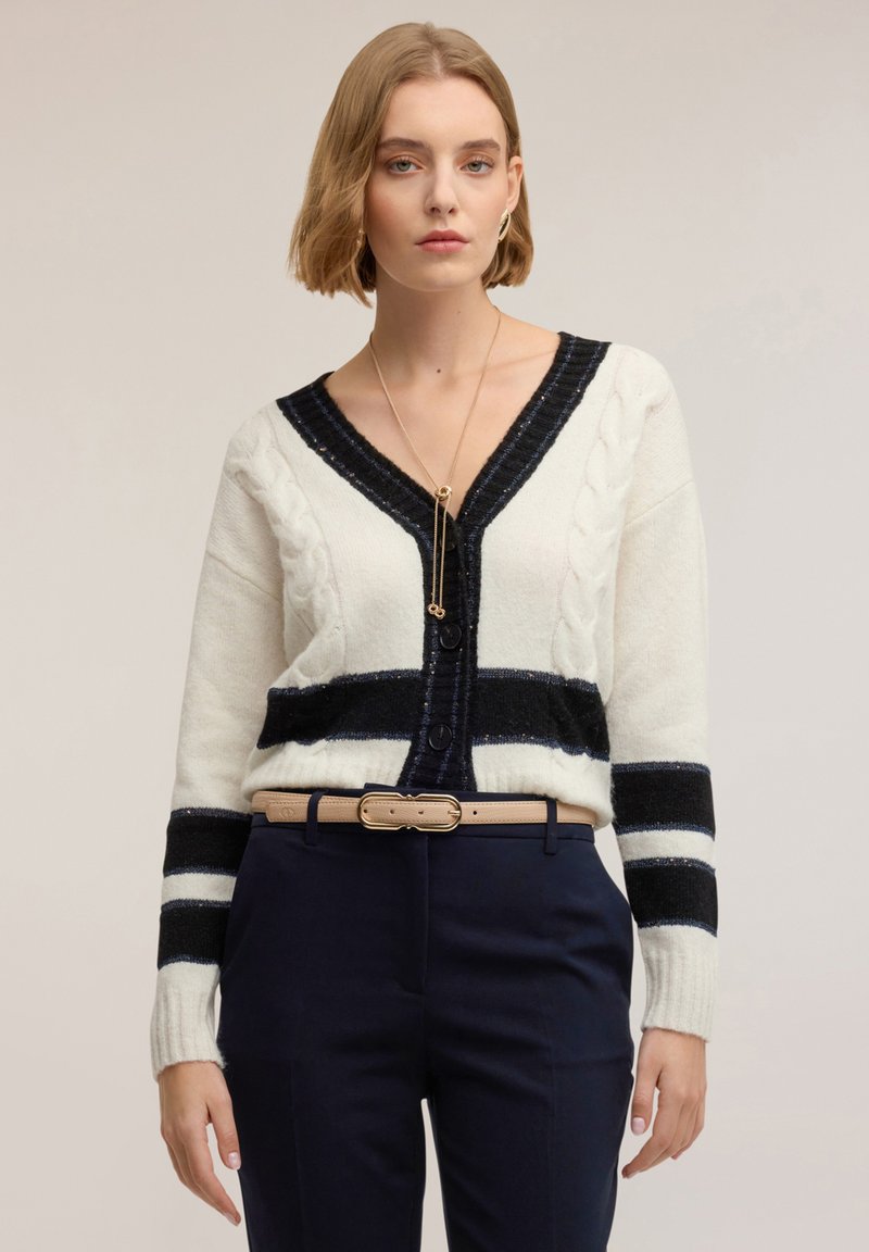 Kremowo-granatowy dzianinowy kardigan z wzorem warkoczowym, dekoltem w serek, paskami i zapięciem na guziki, zestawiony z granatowymi spodniami i beżowym paskiem.