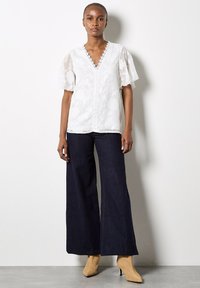 Weiße Bluse mit floralem Muster, V-Ausschnitt und Spitzenbesatz, kombiniert mit weit geschnittenen dunkelblauen Jeans und beigefarbenen Knöchelstiefeln.
