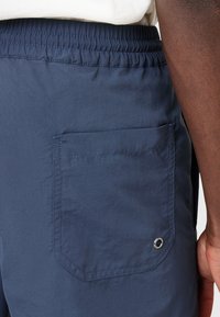 Tummansiniset shortsit joustavalla vyötärönauhasta, takataskussa on kolmion muotoinen läppä ja metallinen silmätyö. Silkkinen pinta.