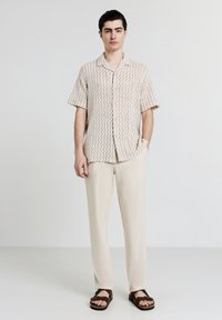 Chemise à manches courtes, boutonnée, avec un motif en zigzag beige et crème ; associée à un pantalon beige clair et des sandales marron.