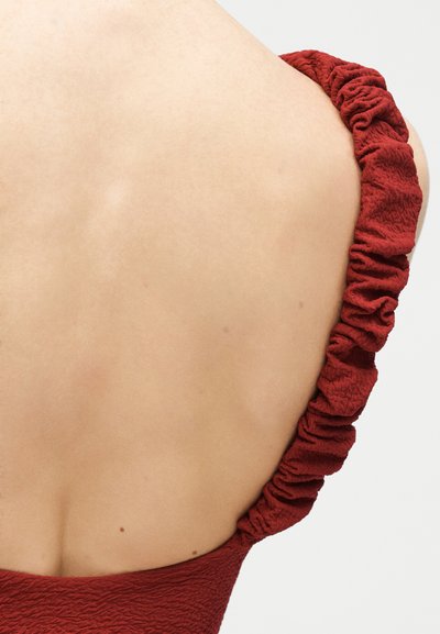 Γυμνή πλάτη με λεία επιδερμίδα που φοράει ένα υφασμάτινο ένδυμα σε βαθύ κόκκινο χρώμα με ραμμένη βολάν ράντα στον δεξί ώμο.