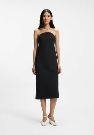 Frau steht in einem trägerlosen schwarzen Midi-Kleid und weißen spitzen High Heels mit Knöchelriemen vor einem schlichten weißen Hintergrund.