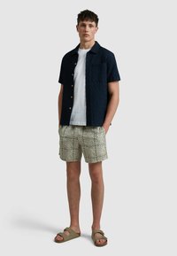 Korte mouw navy shirt met textuur en knopen, wit T-shirt eronder, gestreepte beige shorts, en groene slippers. Eenvoudig ontwerp, casual uitstraling.