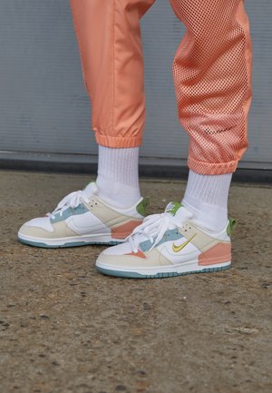 Personne portant des baskets Nike blanches, rose pastel, vertes et bleues avec des chaussettes blanches et un pantalon de jogging couleur pêche avec des panneaux en mesh, debout sur un sol en béton.