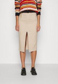 Jupe crayon beige avec une fente avant, fabriquée en tissu texturé. Portée avec un pull rayé coloré et des chaussures noires.
