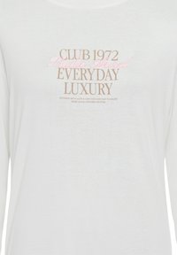 White long-sleeve shirt featuring text in beige and pink, with the phrases "CLUB 1972," "Studio Aure," and "EVERYDAY LUXURY" in various fonts.