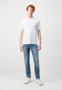 Witte katoenen T-shirt met lichtgrijze tekst, gecombineerd met lichtblauwe spijkerbroek. Model draagt witte sneakers en staat tegen een effen achtergrond.