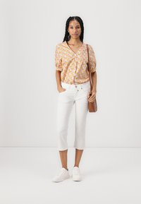 Blusa corta a maniche corte floreale nei toni giallo, rosa e verde, abbinata a pantaloni bianchi a vita alta e sneakers bianche; borsa a tracolla marrone.