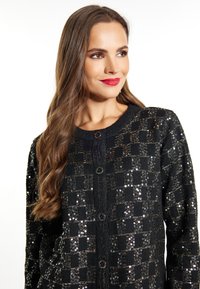 Cardigan negru cu un model de paiete în carouri, guler rotund și trei butoane mari pe față. Textură moale de tricot.