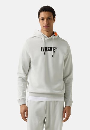 Junger Mann trägt einen hellgrauen Hoodie mit dem Schriftzug „FIRE & ICE“ und passende Hose, steht vor einem weißen Hintergrund.