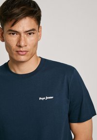 Námořnická modrá bavlněná tričko s kulatým výstřihem a krátkými rukávy, s bílým logem "Pepe Jeans" na levé straně hrudi.