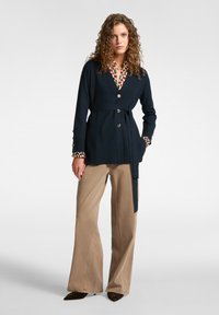 Cardigan navy de estilo envolvente con cintura ajustable, que presenta botones dorados y acentos de puño con patrones, combinado con pantalones de campana beiges.