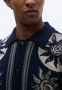 Μπλε ζακάρ  πόλο με μπεζ διακοσμητικές λεπτομέρειες, με κούμπωμα με κουμπιά, ριμπ γιακά και λεπτομερή γραφικό σχέδιο στο ύφασμα.