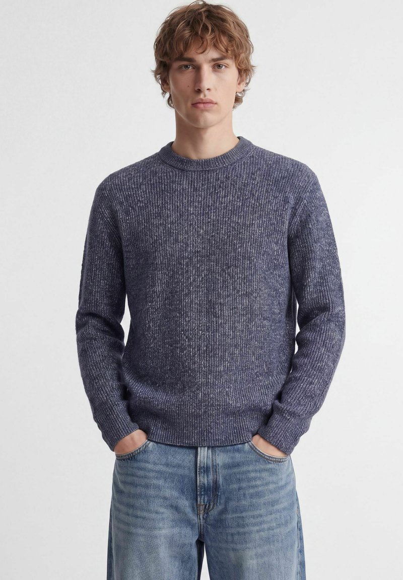 Jeune homme portant un pull tricoté bleu texturé, mains dans les poches, et un jean bleu clair, debout devant un fond blanc uni.