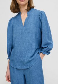 Top blu textured con scollatura a V, spalle arricciate e maniche a sbuffo. Realizzato in tessuto leggero, presenta una vestibilità rilassata.