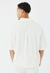 Camisa blanca de manga corta con bordado de cactus en relieve, tela texturizada y corte holgado. Detalles en el cuello y pliegue en la parte posterior visibles.