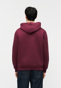 Άνδρας με κοντά σκούρα μαλλιά, ντυμένος με φούτερ μπορντό και σκούρο μπλε τζιν, στέκεται με την πλάτη προς το μέρος μας μπροστά σε λευκό φόντο.