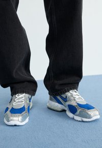 Des chaussures de sport argentées et bleues présentent une tige en mesh, des accents synthétiques et une semelle texturée. Elles sont portées avec un pantalon en denim noir.