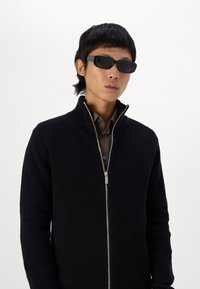 Black zip-up knit sweater with high collar, paired with a grey button-up shirt. Model wears angular black sunglasses. Plain white background.