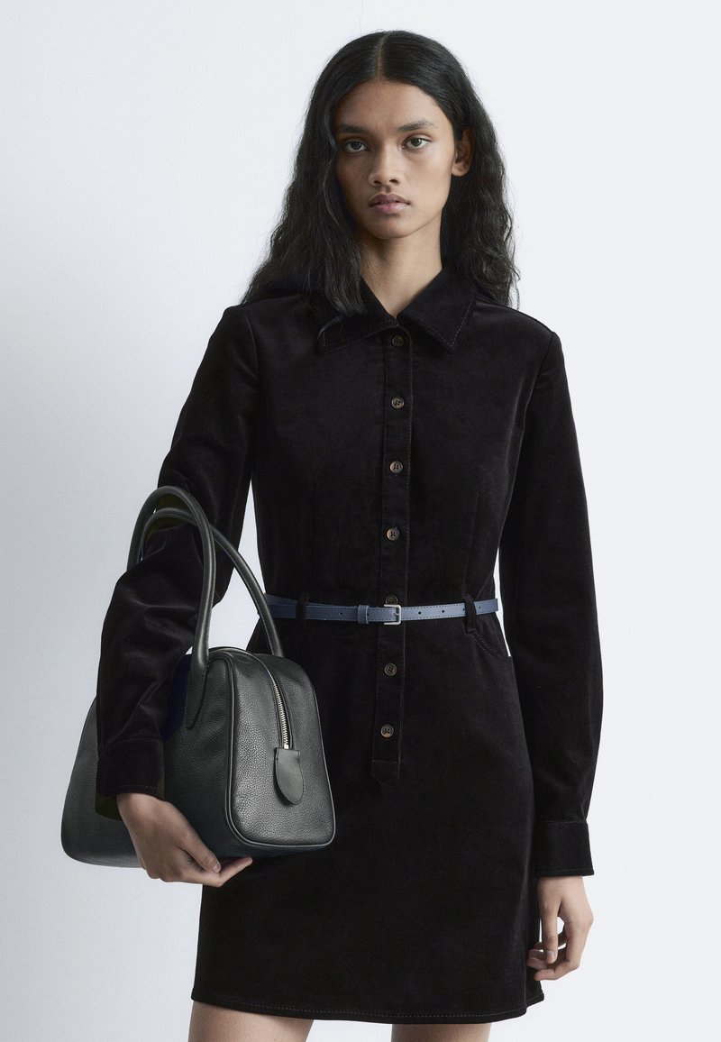Schwarzes Corduroykleid mit Kragen und Knopfleiste, kombiniert mit einer strukturierten schwarzen Ledertasche mit Reißverschlussfach und geschwungenen Griffen.