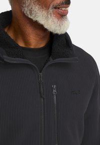 Giacca in pile a coste nera con colletto alto, dotata di un rivestimento morbido e testurizzato e due zip sul davanti. Logo Jack Wolfskin visibile.