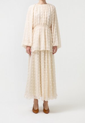 Mujer vistiendo un vestido midi crema de manga larga con lunares texturizados y zapatos marrones de tacón, de pie contra un fondo blanco liso.